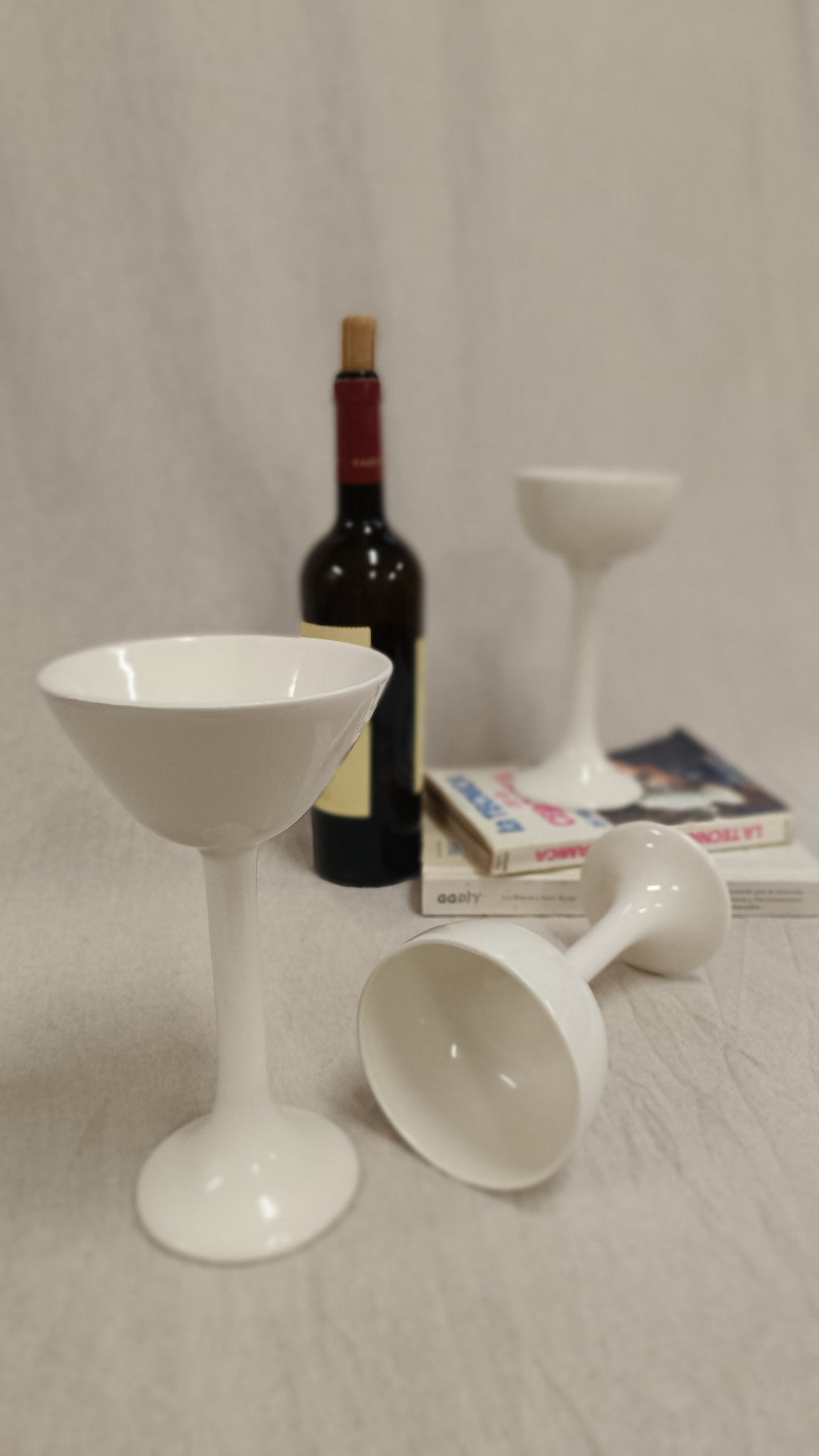 Tres copas de cerámica blanca de Vicente Rivera con diseño redondeado dispuestas junto a una botella de vino y libros.