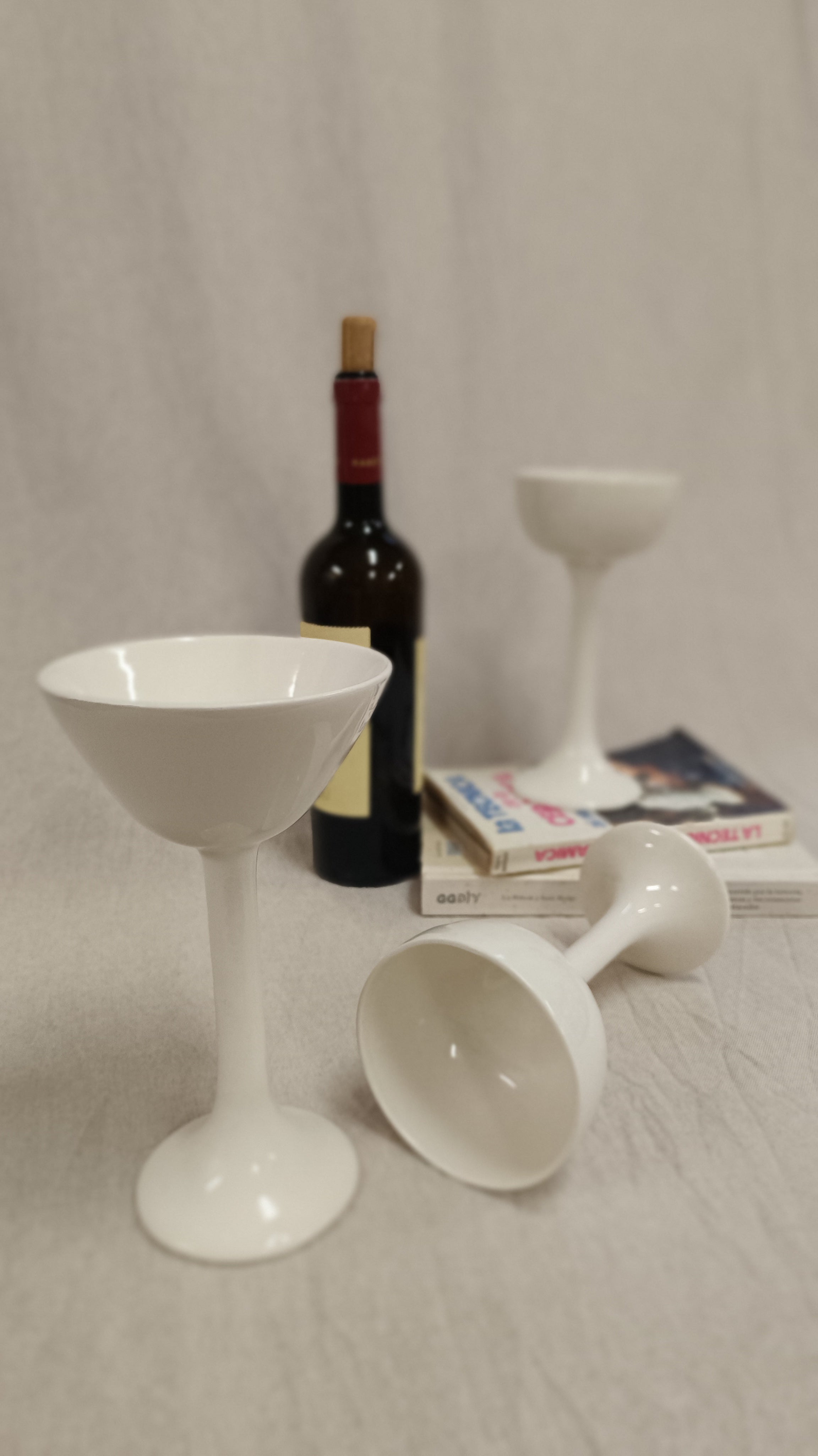 Tres copas de cerámica blanca de Vicente Rivera con diseño redondeado dispuestas junto a una botella de vino y libros.