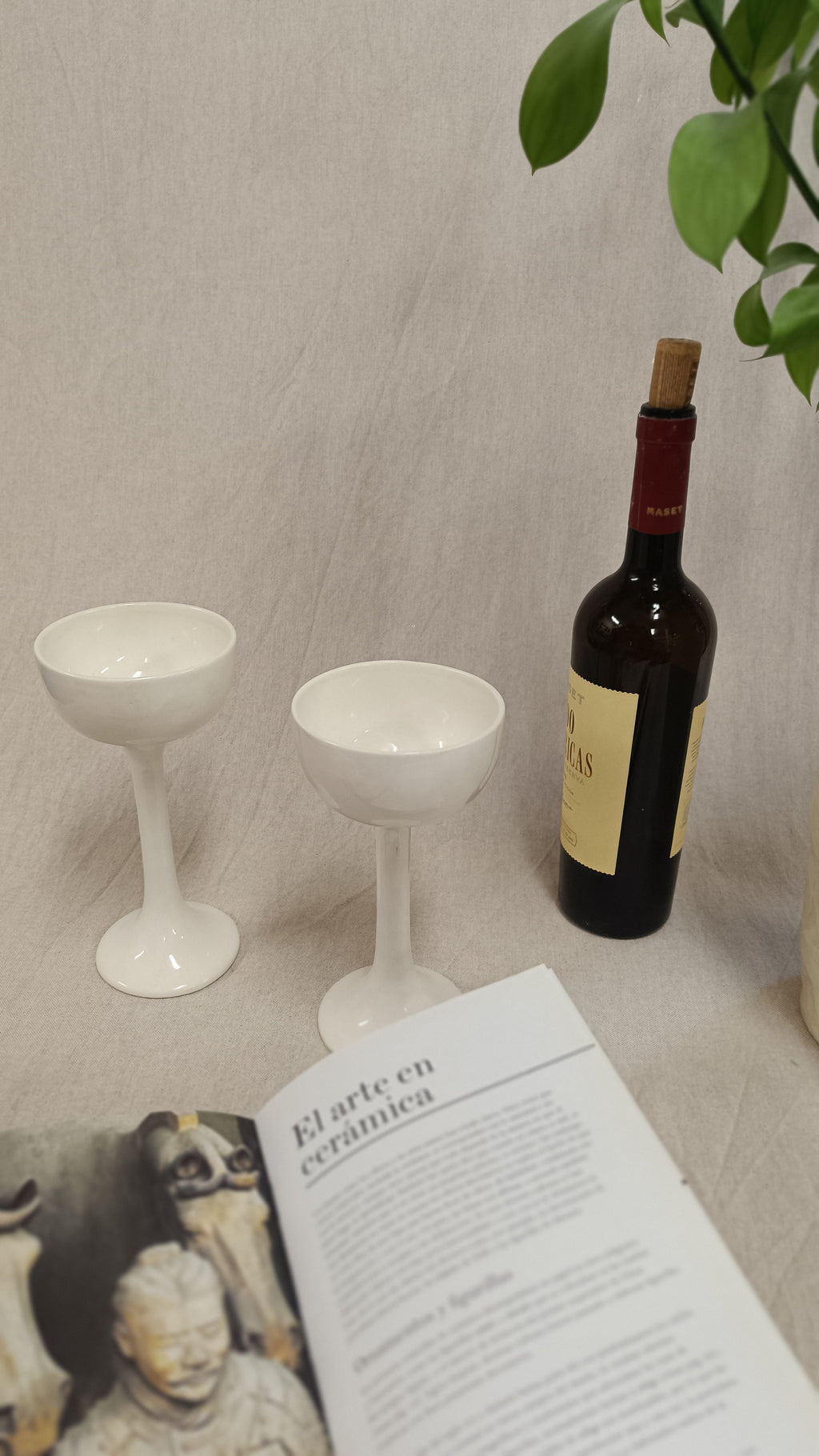 Dos copas blancas de Vicente Rivera integradas en un bodegón con un libro abierto sobre el arte en cerámica y una planta.
