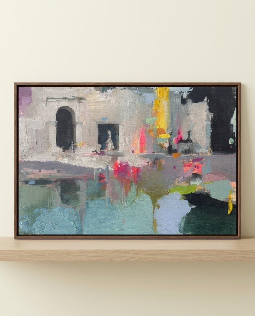 Cuadro enmarcado de la pintura "Jardines de la Memoria" apoyado sobre una repisa de madera clara. La obra abstracta representa ruinas griegas bajo un cielo azul con pinceladas vibrantes de rosa neón y amarillo. Fotografía de decoración con luz natural y cortina blanca.