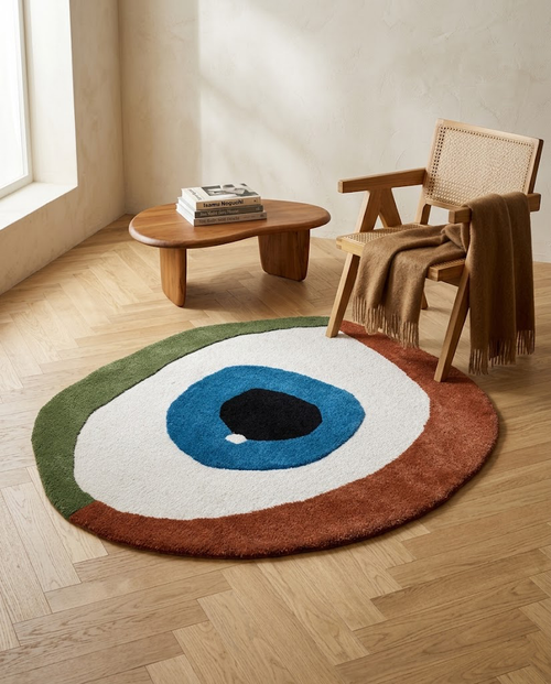 Alfombra de forma orgánica modelo Charm con diseño de ojo azul y verde en un rincón de lectura con silla de rejilla y mesa de madera.