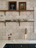 Vista frontal de un elegante frente de cocina revestido con azulejos artesanales blanco roto, con detalles de orlas de esquina y motivos centrales en azul cobalto, sobre una encimera de mármol y estantes de madera con vajilla de vidrio.