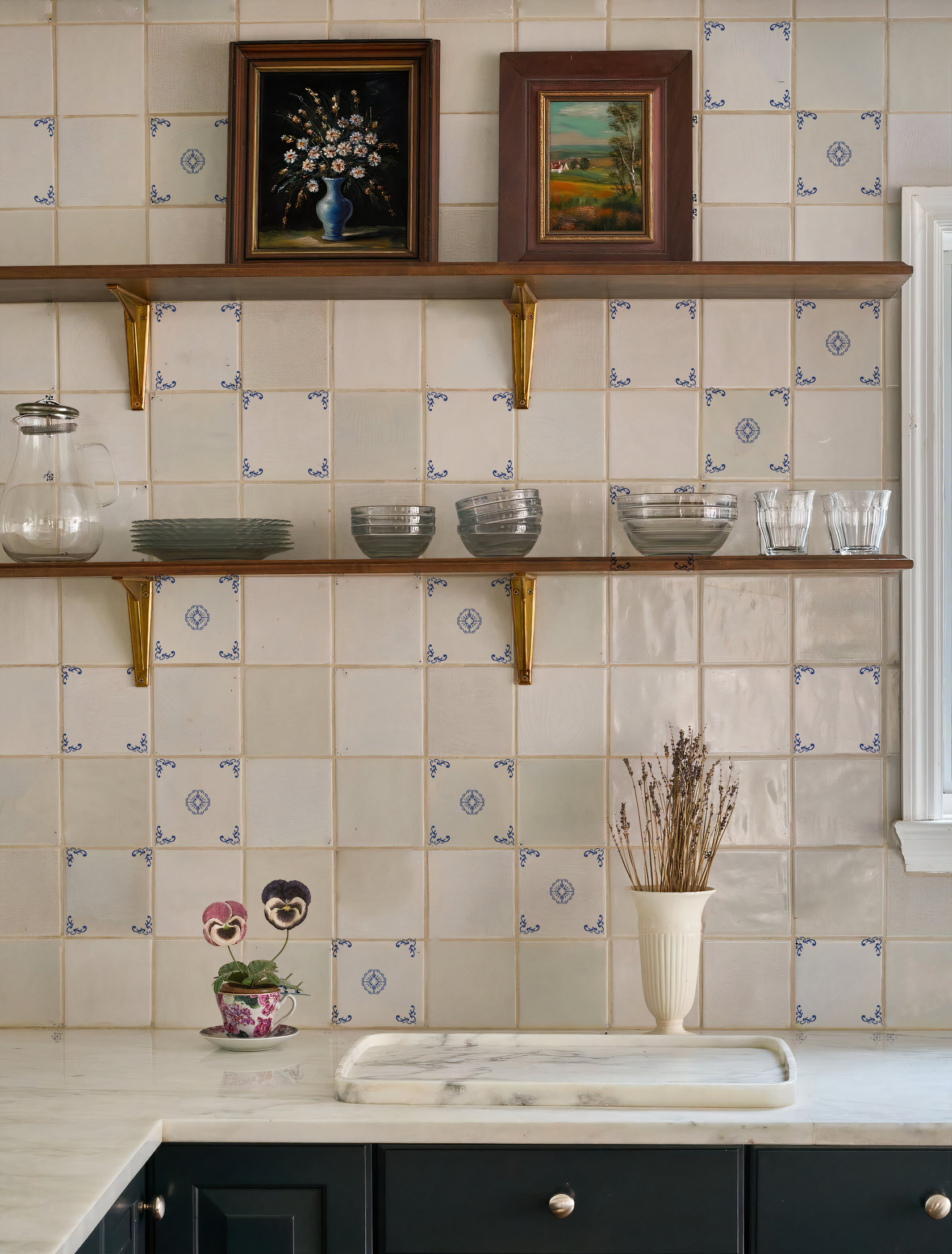 Vista frontal de un elegante frente de cocina revestido con azulejos artesanales blanco roto, con detalles de orlas de esquina y motivos centrales en azul cobalto, sobre una encimera de mármol y estantes de madera con vajilla de vidrio.