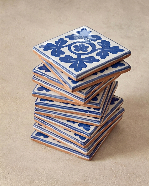 Vista en ángulo de una torre desalineada de azulejos cerámicos artesanales de estilo medieval. El azulejo superior muestra en detalle el patrón "Trébol Medieval" pintado a mano en azul sobre esmalte blanco brillante. Se aprecian las imperfecciones rústicas de la arcilla cocida en los bordes.