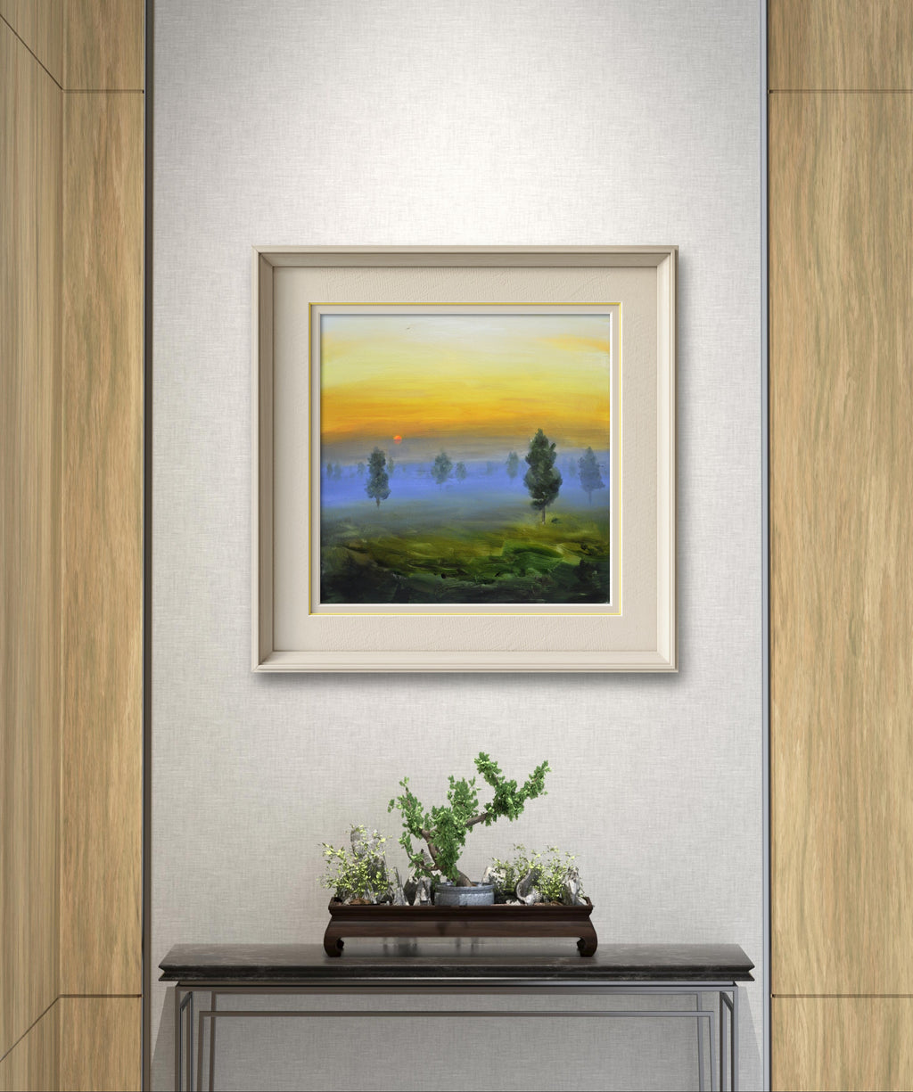 Simulación decorativa de la obra "Amanecer y niebla" enmarcada en una pared minimalista sobre un mueble con un bonsái.