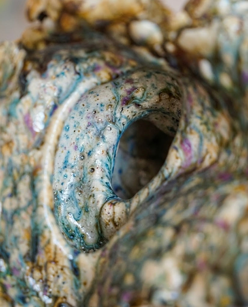 Primer plano macro del interior curvo de una caracola de cerámica de autor, mostrando el efecto poroso y brillante del esmalte reactivo en tonos turquesa, violeta y ocre.