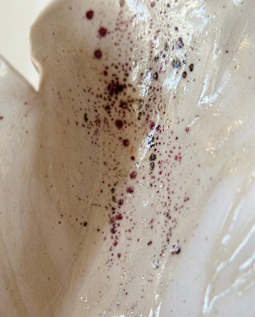 Primer plano macro del interior esmaltado de la escultura Flor Calima, con salpicaduras y motas de pigmento granate y marrón sobre una superficie brillante color crema.