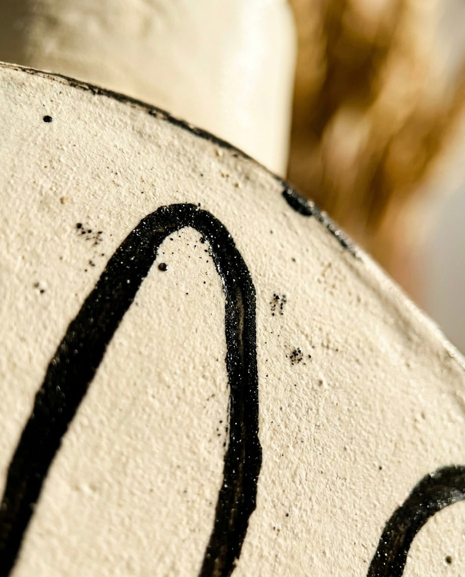 Plano de detalle macro que muestra la textura rugosa de la arcilla blanca y el relieve del trazo negro esmaltado del Jarrón Sorpresa de Magdalena Arnaud.