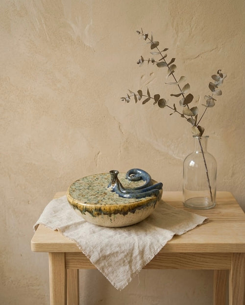 Escultura de cerámica artesanal de autor titulada Auralia Fuentón, colocada sobre un paño de lino en una mesa de madera junto a un jarrón con ramas de eucalipto seco, frente a una pared texturizada beige.
