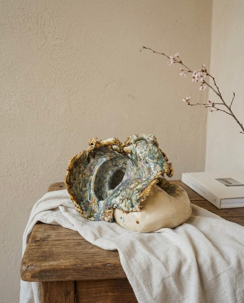 Escultura de cerámica artesanal con forma de caracola marina orgánica y esmaltes reactivos, expuesta sobre un paño de lino en una mesa rústica de madera junto a una rama con flores delicadas.