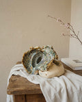 Escultura de cerámica artesanal con forma de caracola marina orgánica y esmaltes reactivos, expuesta sobre un paño de lino en una mesa rústica de madera junto a una rama con flores delicadas.