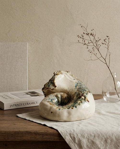 Escultura decorativa de cerámica inspirada en la naturaleza, con base lisa color crema y pliegues texturizados, colocada junto a un libro de diseño Japandi sobre una mesa de madera.
