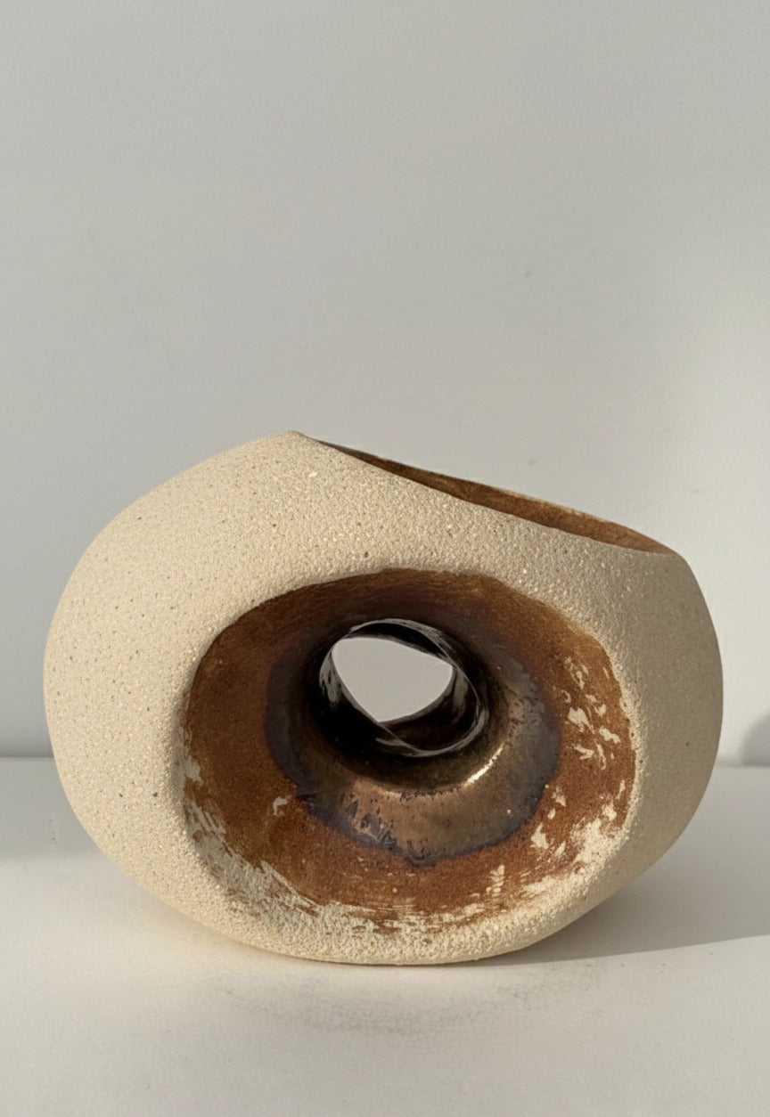 Escultura minimalista de gres "HALO" de Fernanda Salomé, con exterior texturizado crema mate y cavidad interna circular con esmalte reactivo bronce brillante, sobre fondo neutro.