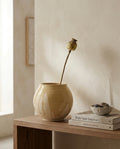 Jarrón esférico de cerámica artesanal de la serie Calima Fuentón en tono ocre arena, sosteniendo una cápsula de amapola seca, sobre un mueble de madera junto a libros de diseño de interiores.