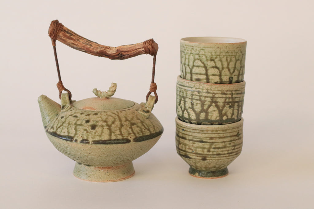 Tetera de cerámica artesanal con texturas verdes y asa de rama natural junto a cuencos apilados.