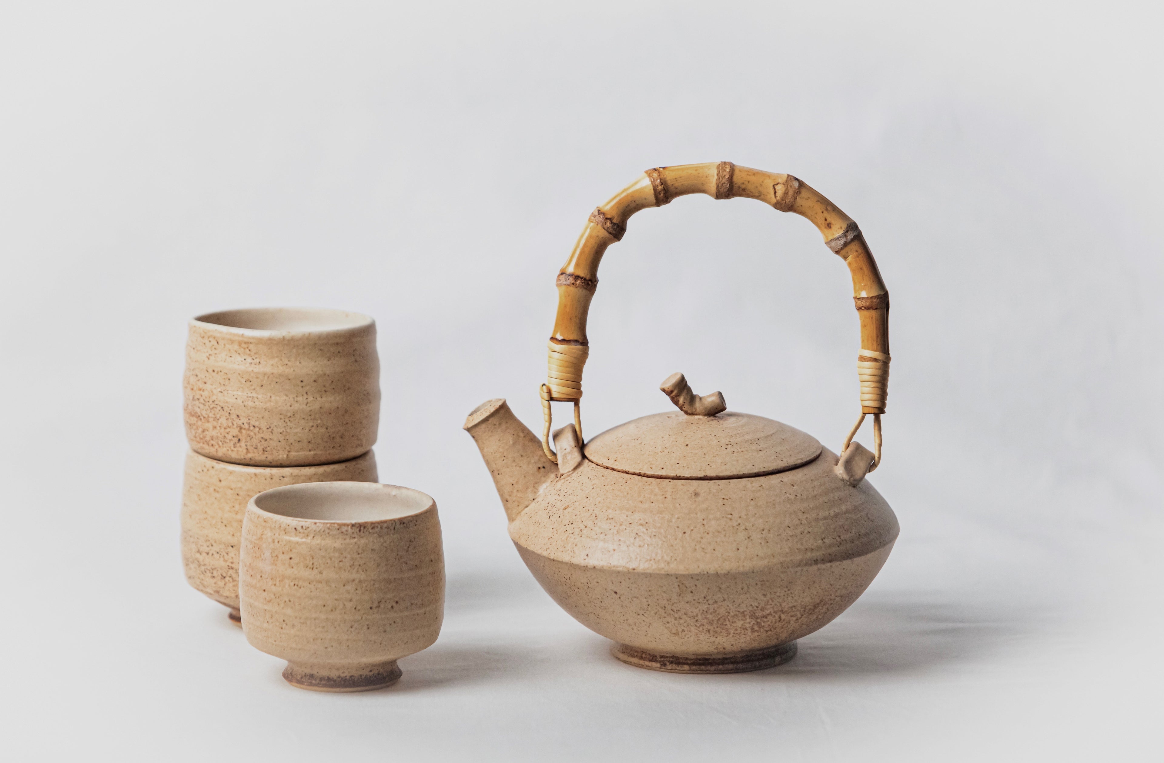 Juego de té de cerámica artesanal en tonos arena con tetera de asa de bambú y vasos a juego sobre fondo blanco.
