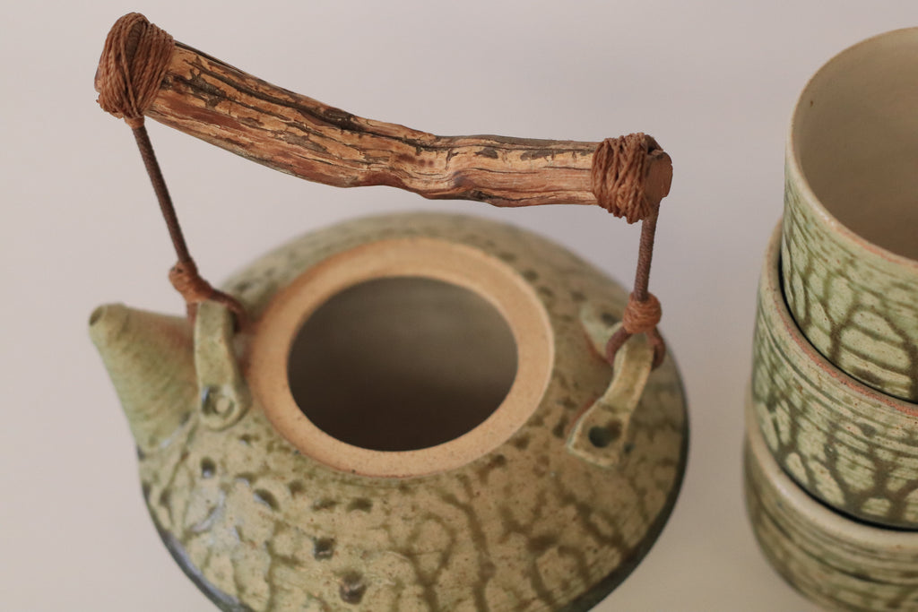 Tetera de cerámica artesanal con textura orgánica en tonos verde musgo y ocre, destacando un asa de rama natural tallada y tres cuencos cilíndricos texturizados a juego.