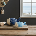 Pareja de esculturas de pez en gres azul (cobalto y celeste) de Vicente Rivera, presentadas sobre madera rústica en un ambiente mediterráneo con vistas al mar, seleccionadas por Sezme Decor.