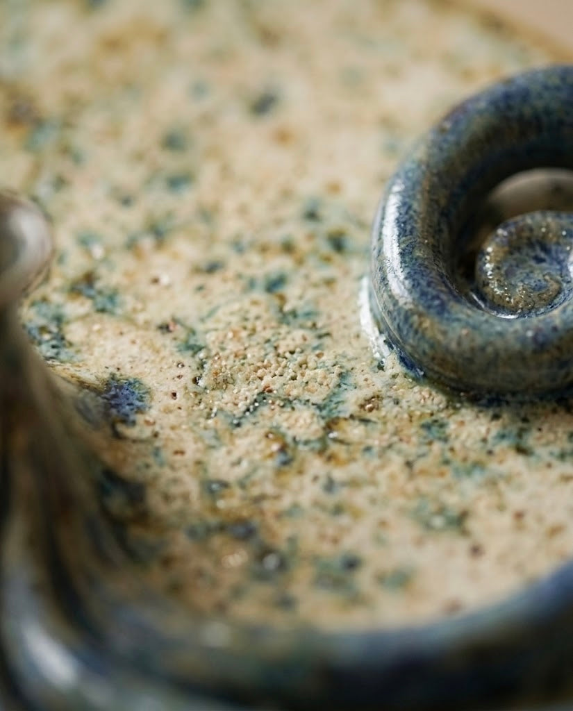 Primer plano macro de la textura de una pieza cerámica, mostrando los detalles del esmalte brillante moteado en tonos tierra y el relieve de un elemento espiral azul cobalto.