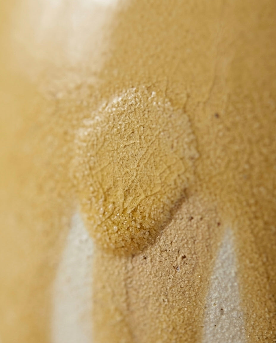 Plano detalle macro que muestra la textura rugosa, porosa y ligeramente craquelada del esmalte cerámico en tonos amarillo mostaza y arena.