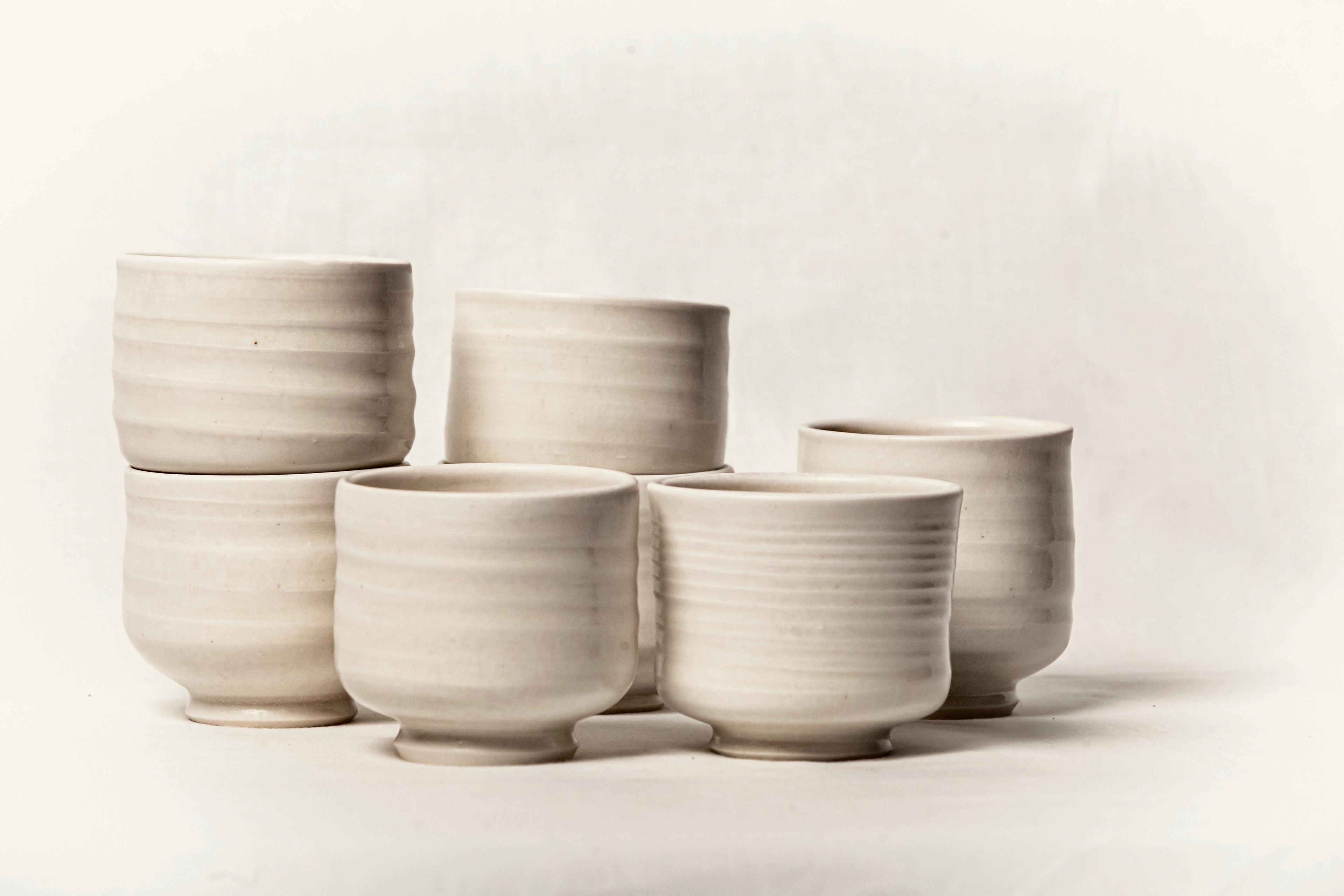 Conjunto de seis vasos de cerámica artesanal en blanco roto, mostrando sus relieves horizontales y formas orgánicas.