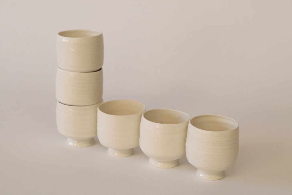 Vasos de cerámica blanca apilados y alineados sobre fondo neutro, resaltando su diseño minimalista y artesanal.